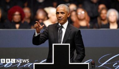 Former US president Barack Obama speaks at the memorial service, wearing a black suit and tie. He's standing on stage with a microphone in front of him and a large cross donning the podium.