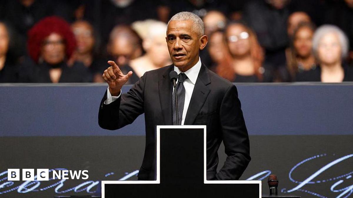 Former US president Barack Obama speaks at the memorial service, wearing a black suit and tie. He's standing on stage with a microphone in front of him and a large cross donning the podium.