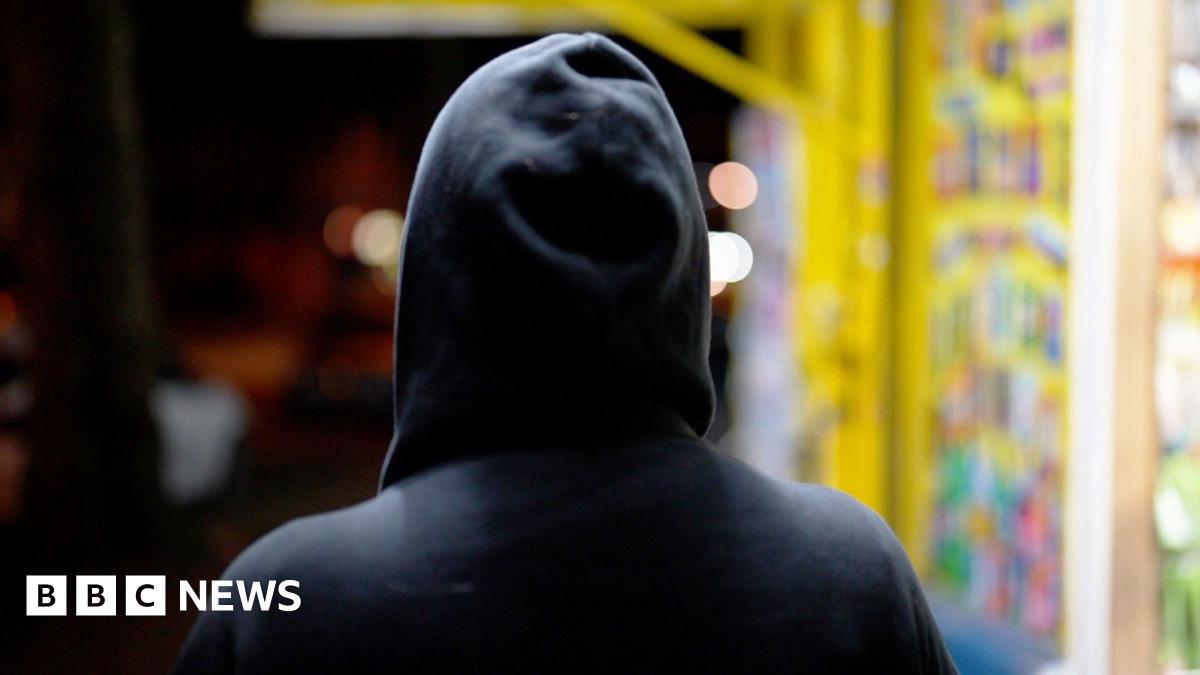 A person in a dark hoodie stands with their back to the camera, facing a brightly lit shop window filled with colourful items. The background is blurred with warm lights, creating a contrast between the dark figure and the vibrant display.