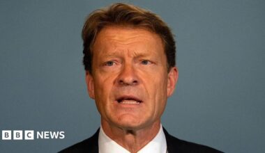 Richard Tice giving a speech. Tice has short brown hair and wears a black suit jacket, white shirt, and a black jacket. mall microphones positioned in front of them on the table.