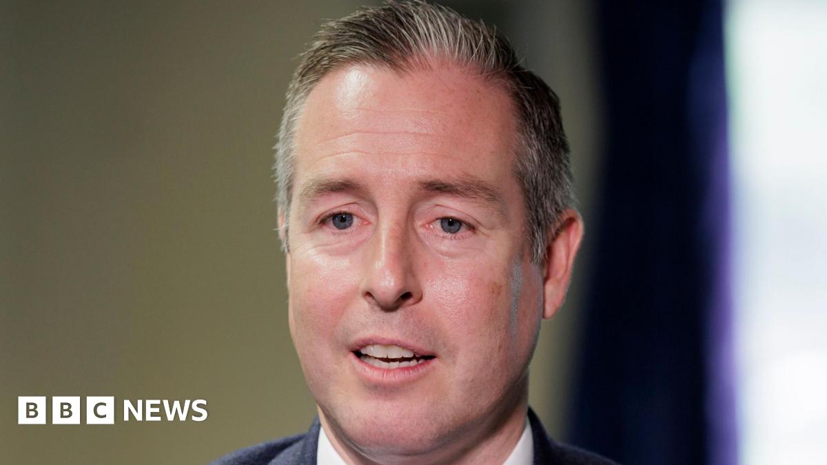 A man with short cropped dark and grey hair is seen head on, the tops of his shoulders are visible in the image. He has his mouth slightly open, as if he is in the middle of speaking. He is wearing a dark jacket and a white shirt. The background is blurred but it appears to be an indoor setting.