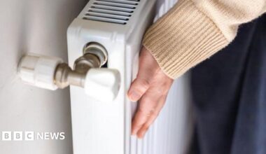 A woman touches a radiator in a house to check how warm it is. She is wearing a beige jumper. The close-up picture shows her hand and sleeve.