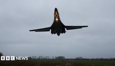 A large bomber aircraft is pictured with a bright light shining as it approaches a runway at an airbase.