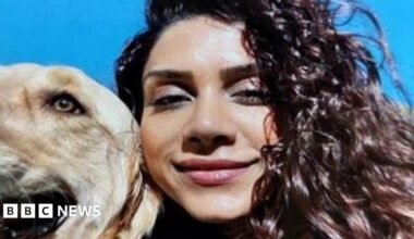 A close up selfie of Paria with a blonde golden retriever dog on a sunny day. She has brown curly hair and is smiling.