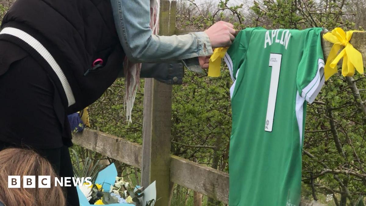 A green shirt with Amelia's name and the number 1 hangs from a fence whilst a woman signs it.