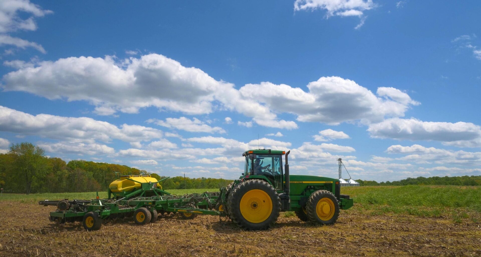 A green and yellow tractor pulls farming equipment across a field under a blue sky with scattered clouds.