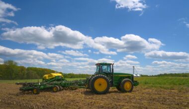 A green and yellow tractor pulls farming equipment across a field under a blue sky with scattered clouds.