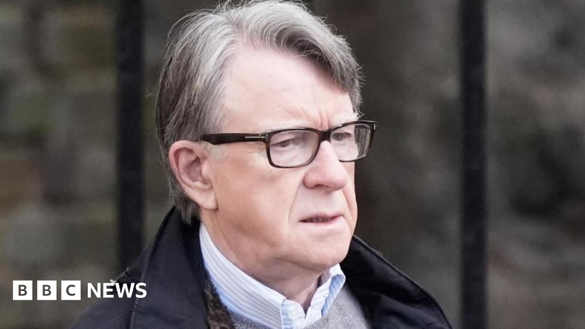 Lord Mandelson wearing glasses, a dark coloured coat, a grey jumper and a shirt.