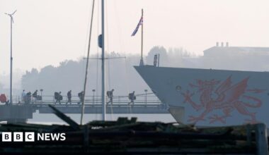 Crew members board the HMS Dragon during ammunitioning operations. A red dragon is painted on the side of the grey ship.