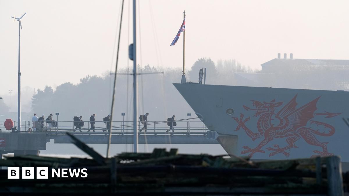 Crew members board the HMS Dragon during ammunitioning operations. A red dragon is painted on the side of the grey ship.