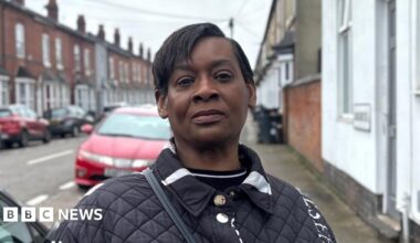 Lorraine Boyce is an elderly woman. She is sitting in her lounge on a brown chair. We can see a bookcase behind her. There is a mug on the table next to her. She is wearing clear thin-rimmed glasses and a dark blue jumper.