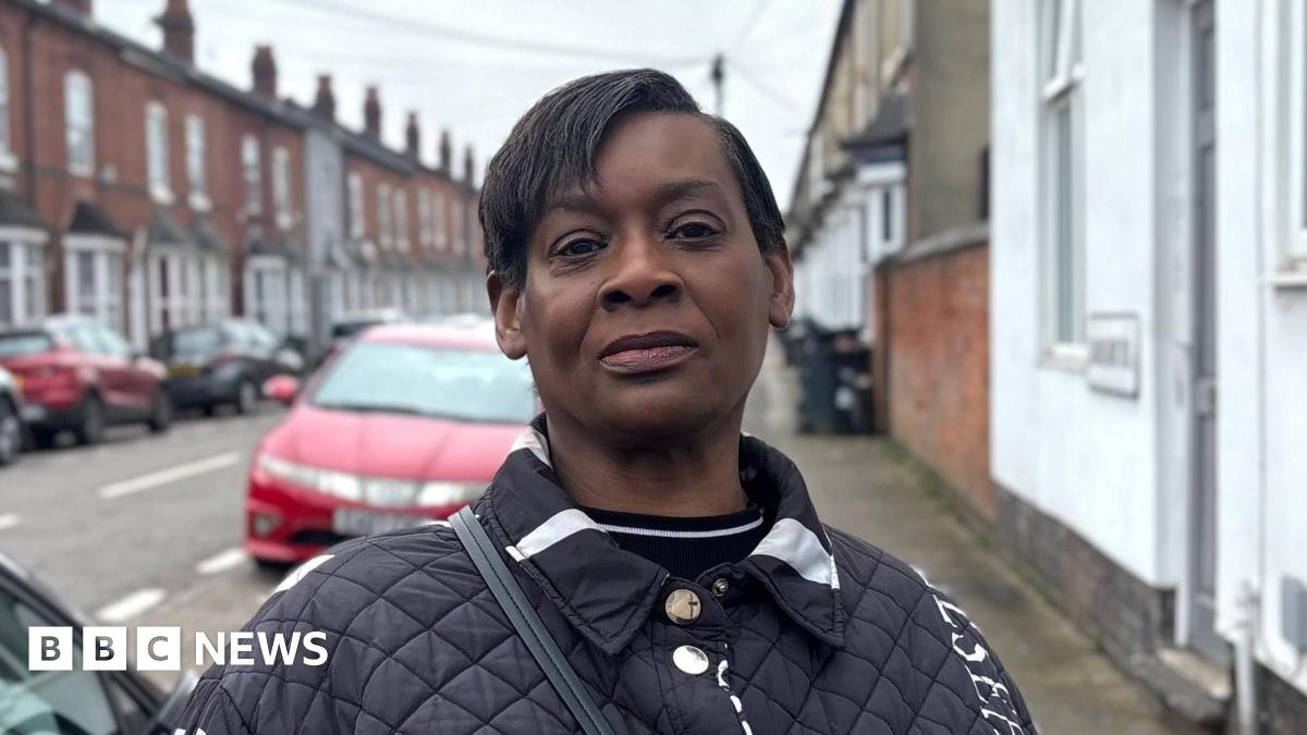 Lorraine Boyce is an elderly woman. She is sitting in her lounge on a brown chair. We can see a bookcase behind her. There is a mug on the table next to her. She is wearing clear thin-rimmed glasses and a dark blue jumper.