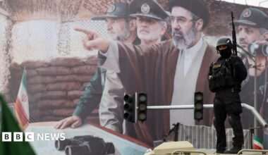 A woman with dark hair and a multiple colour jumper siting on a green sofa inside her living room.