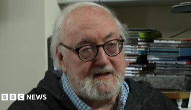 Martin Shipton is wearing a light wash black zip hoody and a blue chequered shirt underneath. He has a beard and is wearing glasses. Behind him are books and portraits.