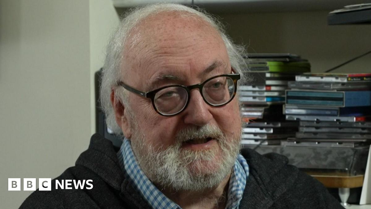 Martin Shipton is wearing a light wash black zip hoody and a blue chequered shirt underneath. He has a beard and is wearing glasses. Behind him are books and portraits.