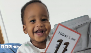 A young child with dark hair wearing a white T-shirt with coloured spots on it is smiling. He is holding a card which reads: "TODAY I AM 11," although the bottom of the card is cut off.