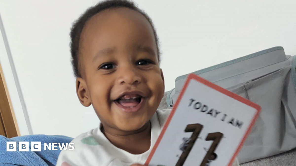 A young child with dark hair wearing a white T-shirt with coloured spots on it is smiling. He is holding a card which reads: "TODAY I AM 11," although the bottom of the card is cut off.