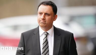 Anas Sarwar, who has short black hair, looks to his right while walking outside. He is frowning. He is wearing a black suit, white shirt and black and silver stripy tie.