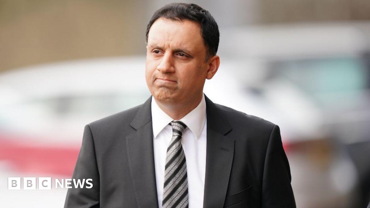 Anas Sarwar, who has short black hair, looks to his right while walking outside. He is frowning. He is wearing a black suit, white shirt and black and silver stripy tie.