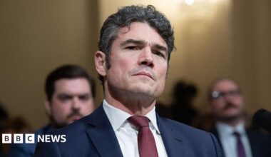 Kent during a committee hearing, wearing a dark blue suit and a red tie, with a serious expression