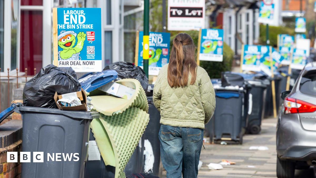 A girl in jeans and green jacket walks past bins overspilling with rubbish on a residential street.