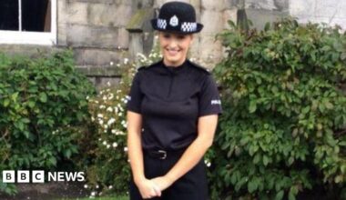 A woman with blonde hair smiling. She is wearing a police uniform and standing in front of green bushes.