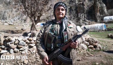 Shaho Bloori, dressed in grey uniform, stands to the right of the picture looking up and to the left in a barren mountainous landscape with a few small trees