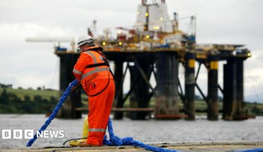 A person wearing orange reflective work gear and a white construction helmet pulls a blue rope while standing on the edge of the water as a mobile offshore drilling unit is transported into the harbour in the Port of Cromarty Firth in Cromarty in 2016.