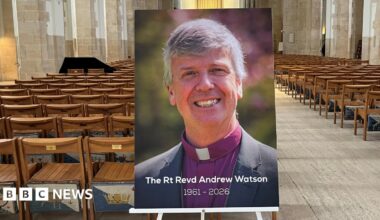 A photo of a bishop propped on a stand, captioned with "The Rt Revd Andrew Watson 1961-2026" at the bottom. It is placed inside a cathedral building lined with seats.