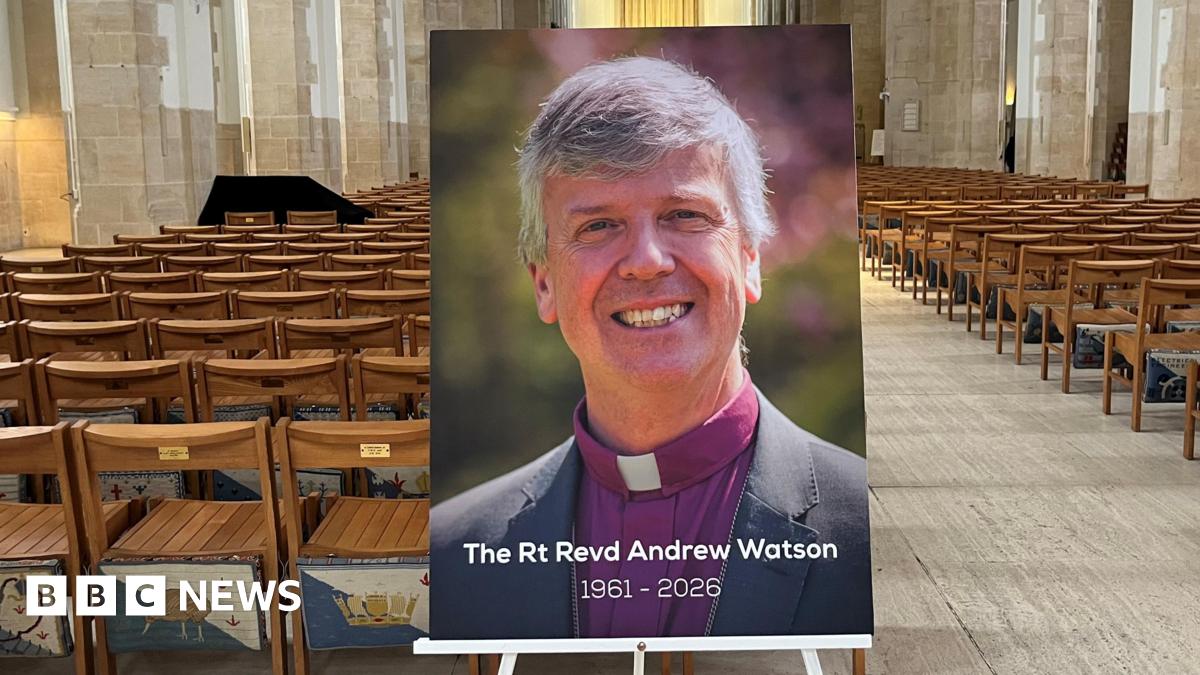 A photo of a bishop propped on a stand, captioned with "The Rt Revd Andrew Watson 1961-2026" at the bottom. It is placed inside a cathedral building lined with seats.