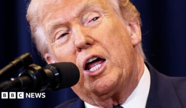 US President Donald Trump speaks into a microphone, his mouth is agape and he is wearing a blue suit jacket and white shirt. Behind him is a dark blue curtain.