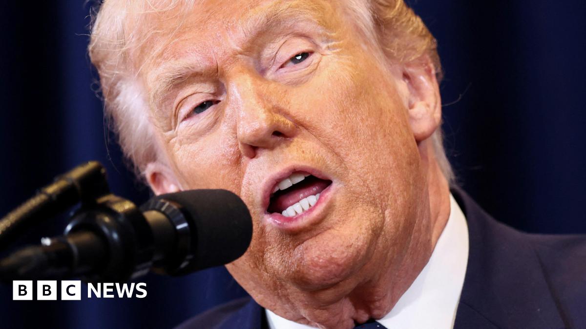 US President Donald Trump speaks into a microphone, his mouth is agape and he is wearing a blue suit jacket and white shirt. Behind him is a dark blue curtain.