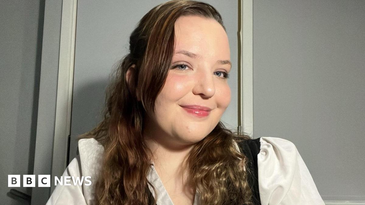 Aeryn Hill smiles at the camera and looks to the side. She has long wavy brown hair and is wearing white