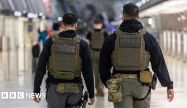 Vik Fielder outside. She is wearing glasses, has curly grey hair and is in a dark grey jacket over a blue T-shirt. She is in front of a stone wall.