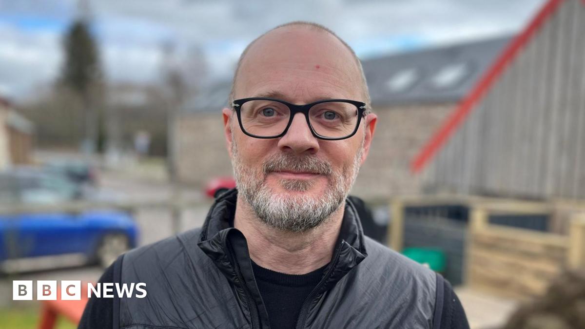 David Watson has grey hair and beard. He is wearing black rimmed glasses and black outdoor clothing. He is standing in a garden area with buildings and a car behind him.