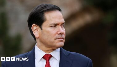 A row of the leaders stood in a line facing forward and smiling. They're on a short stage and are dressed smartly. A fancy old stone building with archways is in the background