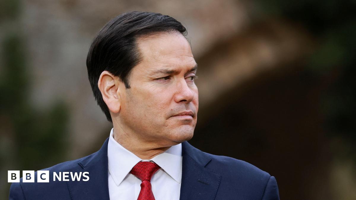 A row of the leaders stood in a line facing forward and smiling. They're on a short stage and are dressed smartly. A fancy old stone building with archways is in the background