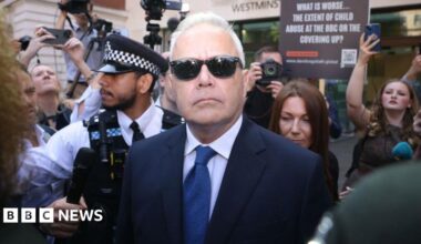 Huw Edwards, wearing a suit and sunglasses, outside court, surrounded by media and protestors