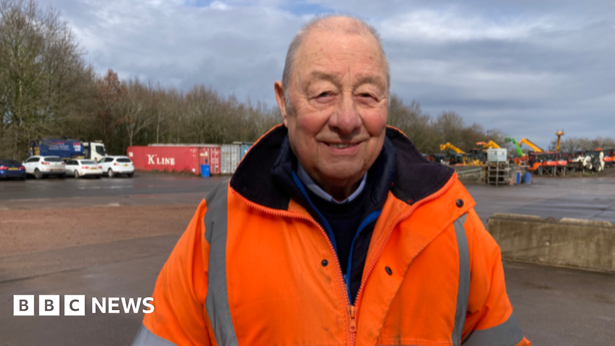 A man in an orange fluorescent jacket smiles in a large carpark with storage containers and other vehicles in the background.