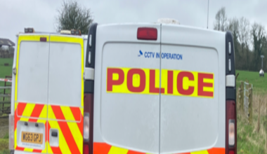 Two police vans on a driveway at Dunkeswell Aerodrome. There is a grass field with a red tent in background.