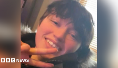 Close up picture of Demi smiling looking at the camera. She has short brown hair and wears a black coat with a fur hood. She is doing a peace pose with her one hand.