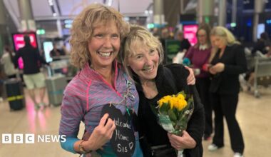 Elaine and Norita standing in an airport terminal, arms around each other and broad smiles on their faces - one is holding a "welcome home sis" sign and the other is holding a bunch of yellow roses