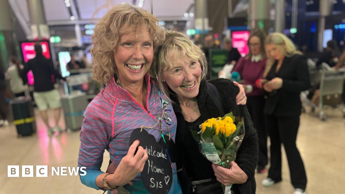Elaine and Norita standing in an airport terminal, arms around each other and broad smiles on their faces - one is holding a "welcome home sis" sign and the other is holding a bunch of yellow roses