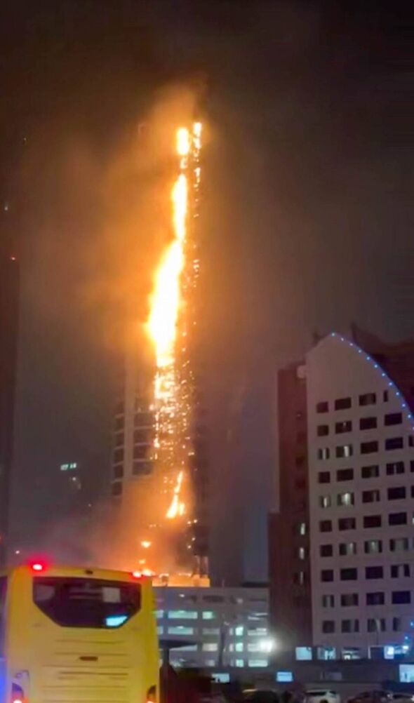 An image depicting a yellow bus parked in front of a building with significant fire damage visible in the background. The scene 