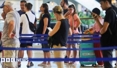 Several people in colourful summer clothes queue around blue barriers at a bright airport forecourt.