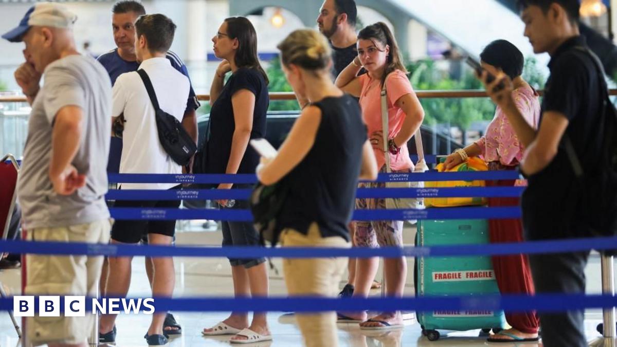 Several people in colourful summer clothes queue around blue barriers at a bright airport forecourt.