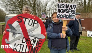 Two women stand in front of other protesters near an RAF Fairford entrance sign. They are holding signs, one is round and in the format of a giant stop sign in red. It features a painted military drone and the words "Stop drone killing". The other sign simply states "US war mongers go home" in black capital letters.