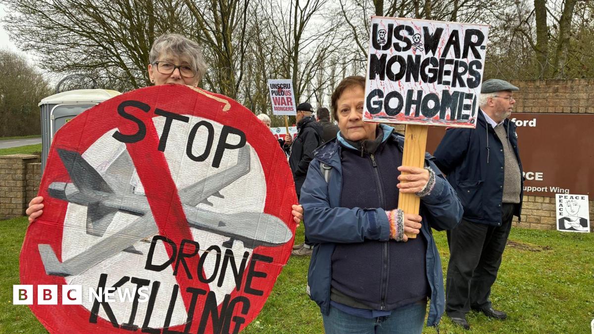 Two women stand in front of other protesters near an RAF Fairford entrance sign. They are holding signs, one is round and in the format of a giant stop sign in red. It features a painted military drone and the words "Stop drone killing". The other sign simply states "US war mongers go home" in black capital letters.