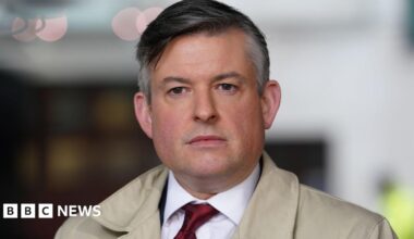Jonathan Ashworth wearing shirt and tie and a cream coat