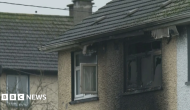 The camera is zoomed in on the top two windows of a light brown house. The window on the right id smashed and is black with burn marks. The area around the window is also stained black. The window to the right is open and relatively untouched by heat.